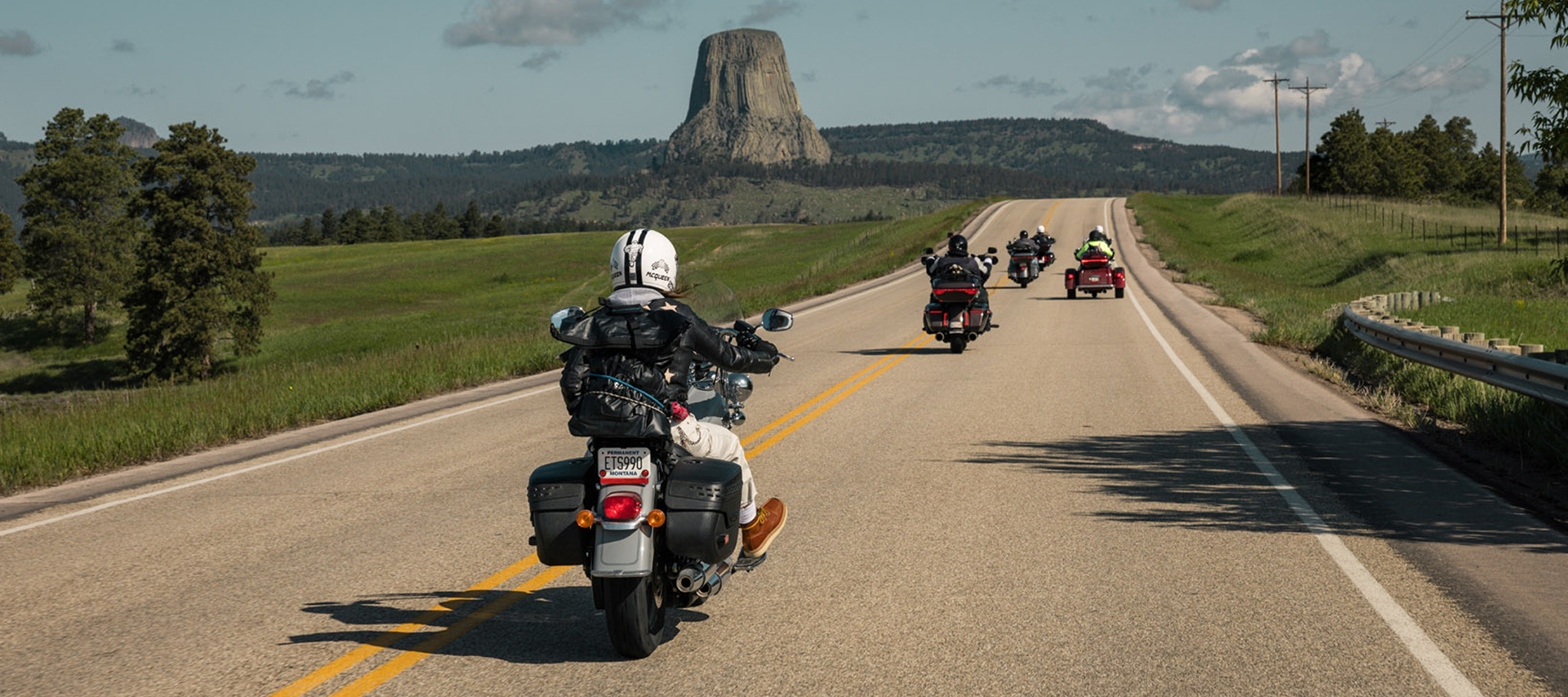 Motorcyclists riding on an open highway toward Devils Tower under a clear sky with green fields