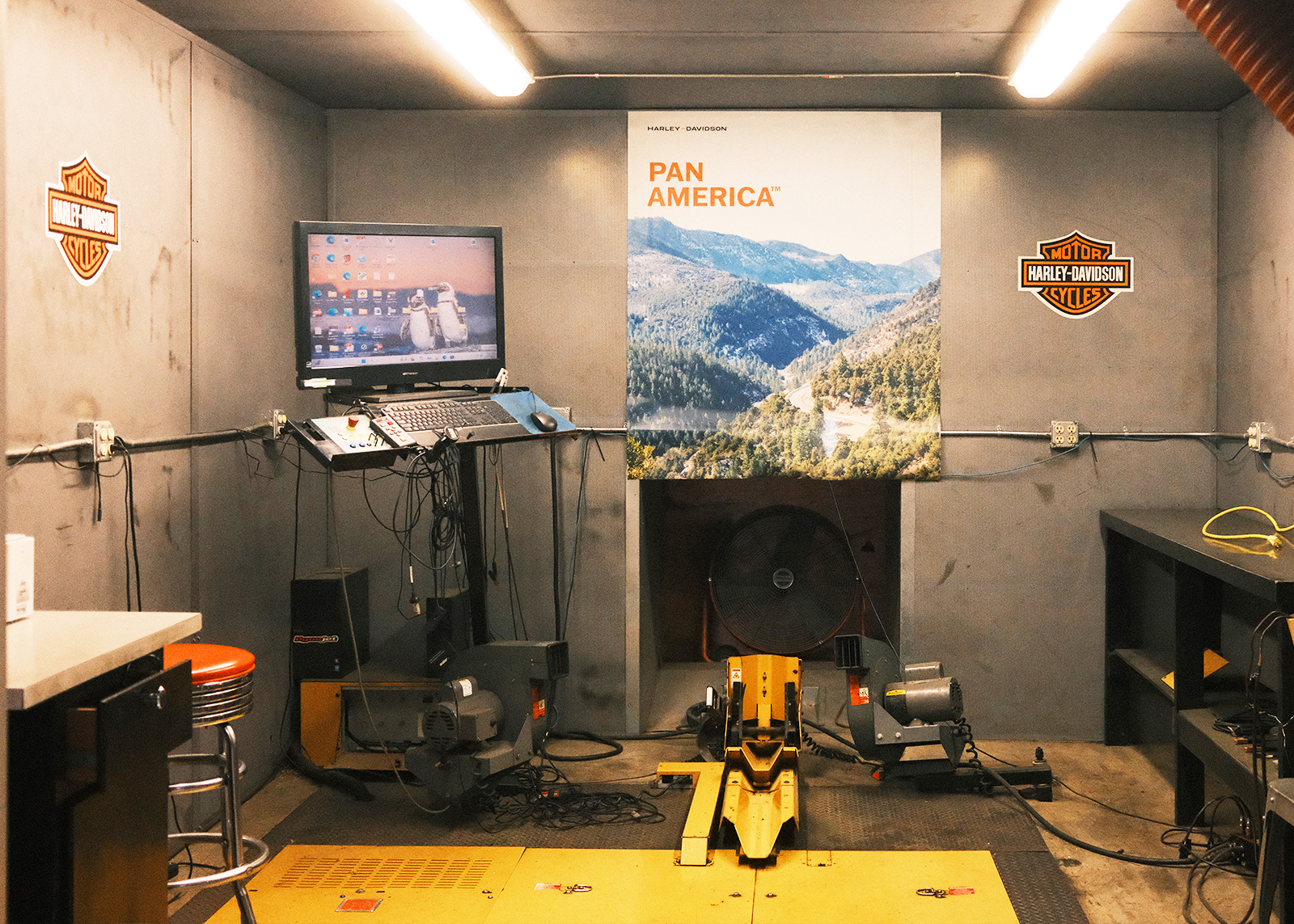 An empty garage bay with a computer, motorcycle stand, and H-D Pan America poster hanging on the wall.
