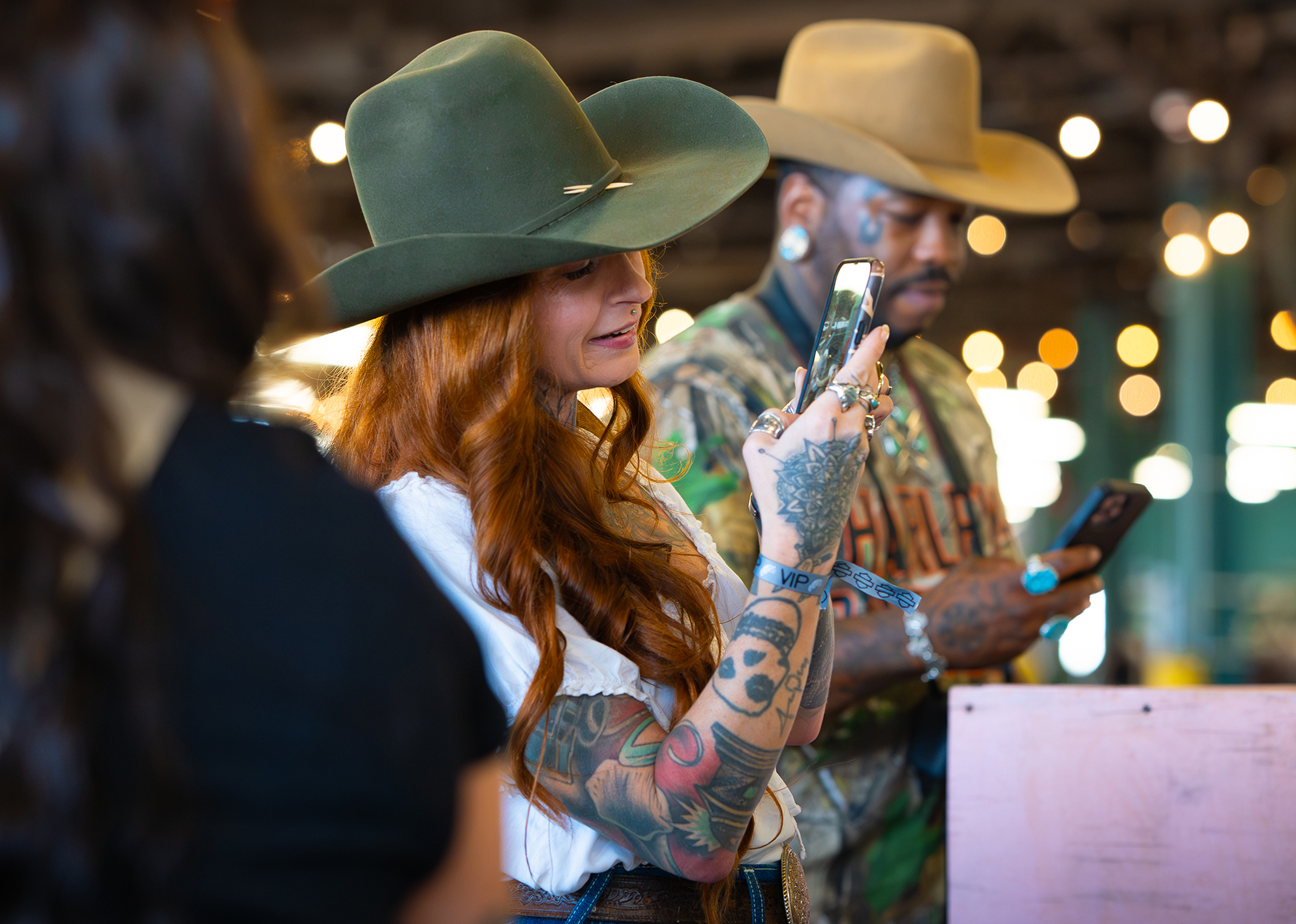 Tattooed woman in a wide-brim hat snaps a photo on her phone as another camo-clad guest checks his.