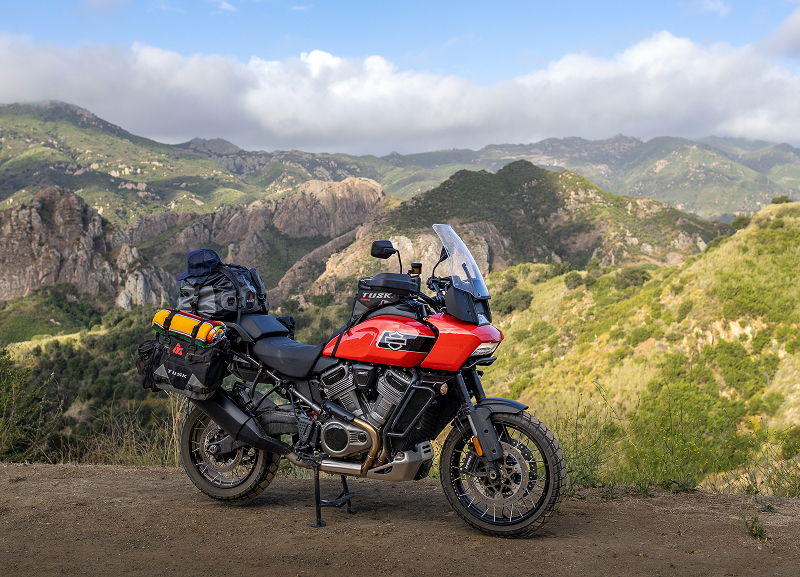 Red H-D Pan America motorcycle with full luggage parked on dirt overlook, mountainous landscape and clouds behind