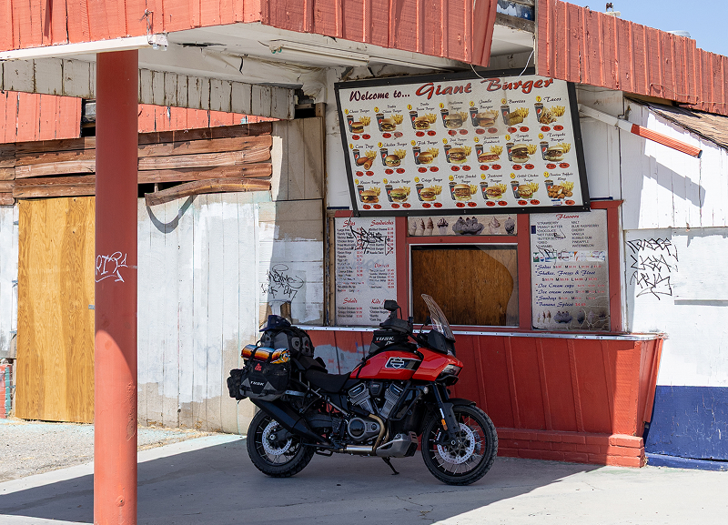 Red H-D Pan America motorcycle loaded with gear parked at roadside food stand with menu board and weathered building