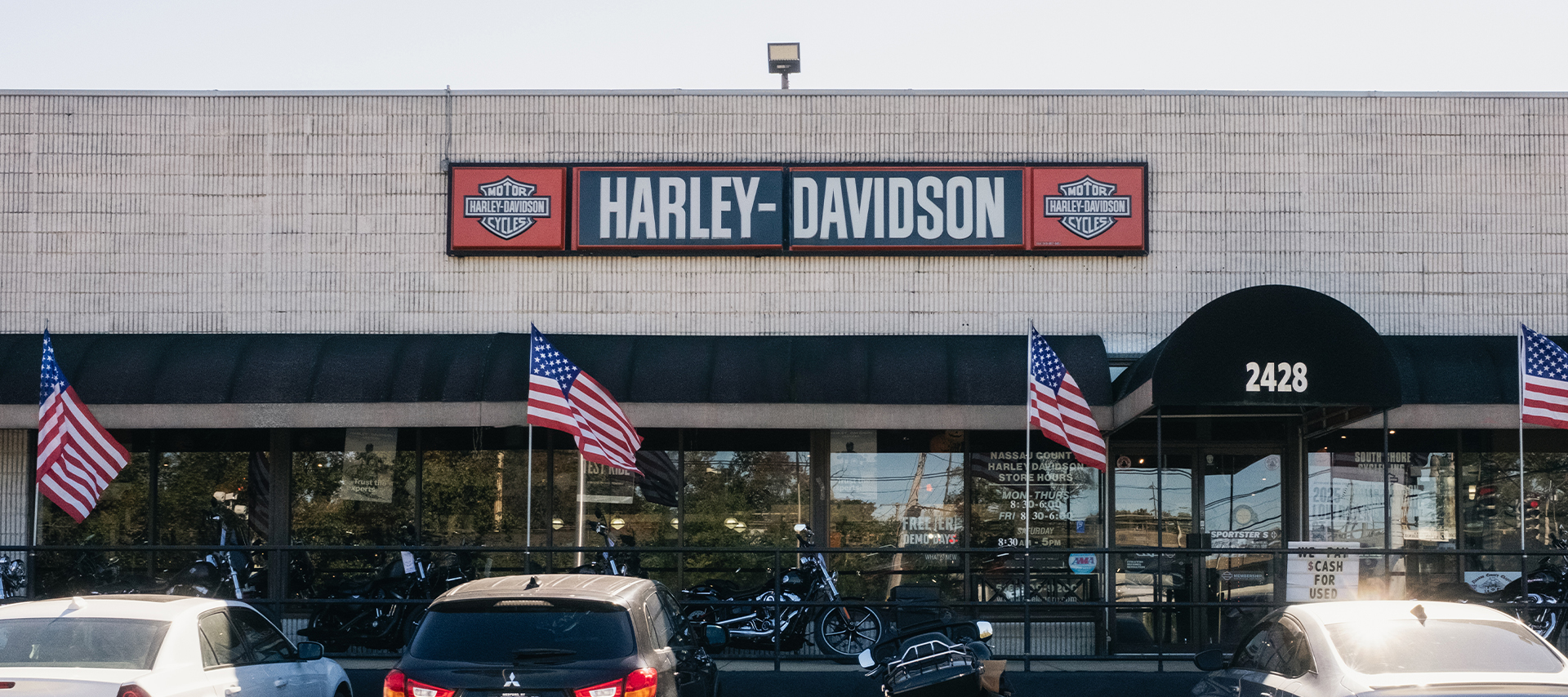 Exterior of Harley-Davidson of Nassau County dealership with parked vehicles and American flags lining the entrance.
