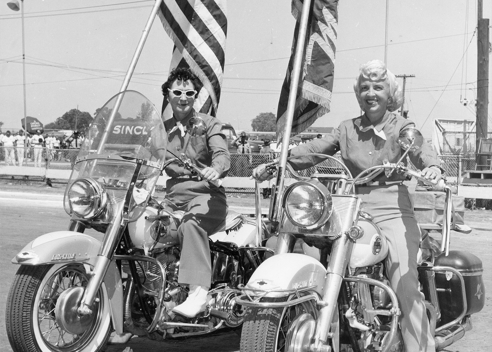 A vintage photo of two Motor Maids sitting on their motorcycles at a rally, one displaying an American flag and the other displaying a Motor Maids flag.