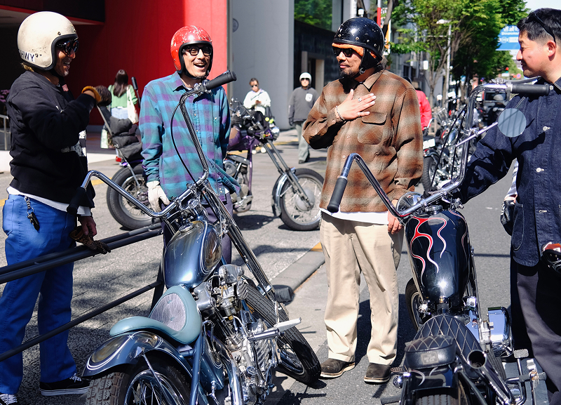 Group of riders in helmets standing beside parked custom motorcycles on a city street, talking near buildings and traffic