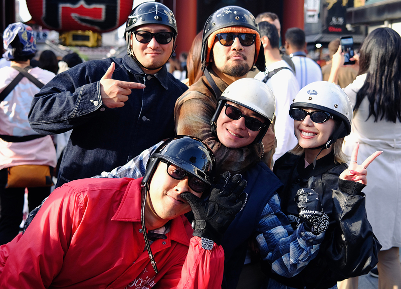 Group of helmeted riders gathered closely in a busy outdoor setting with buildings and a large lantern visible in the background