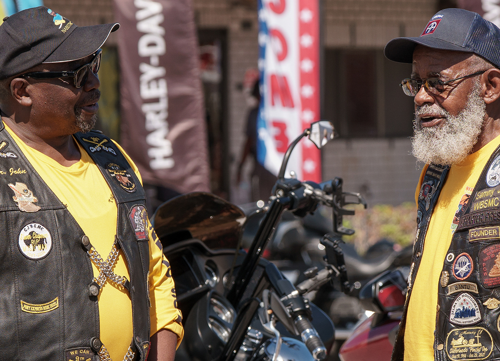 Two people in matching long sleeve yellow shirts and motorcycle vests with patches stands beside a parked bike