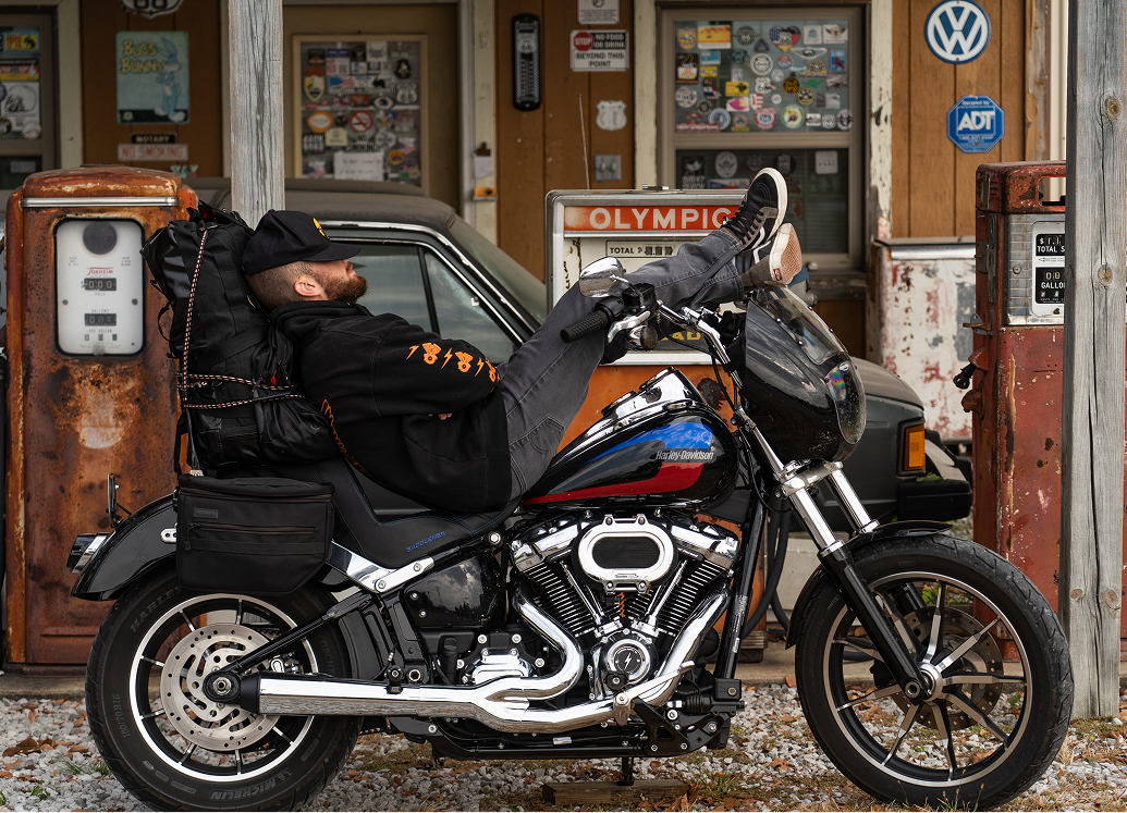 Motorcycle parked in front of a vintage gas station, with a person reclining on the seat and gear strapped on