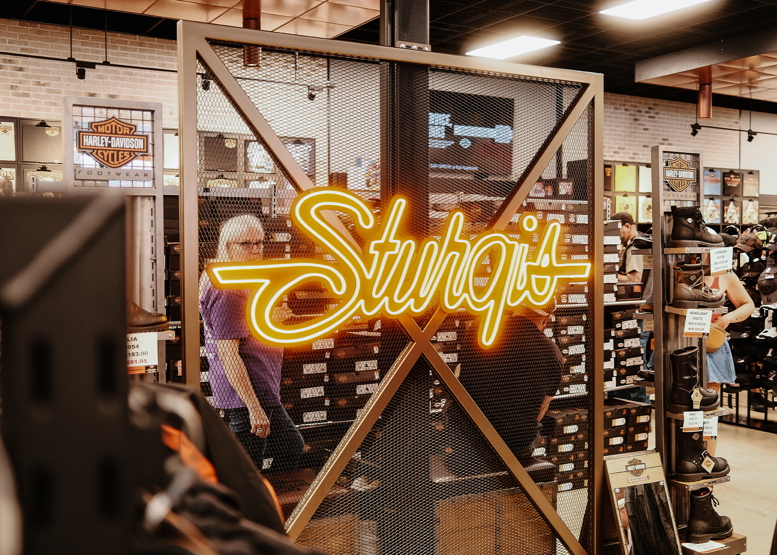 Neon orange “Sturgis” sign inside the Sturgis Harley-Davidson store with people browsing boots and apparel.