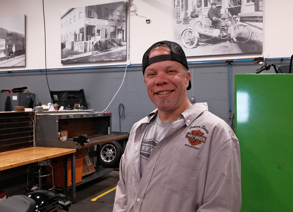 Service technician wearing backwards hat and smiling standing near workbench, tools, and motorcycle equipment
