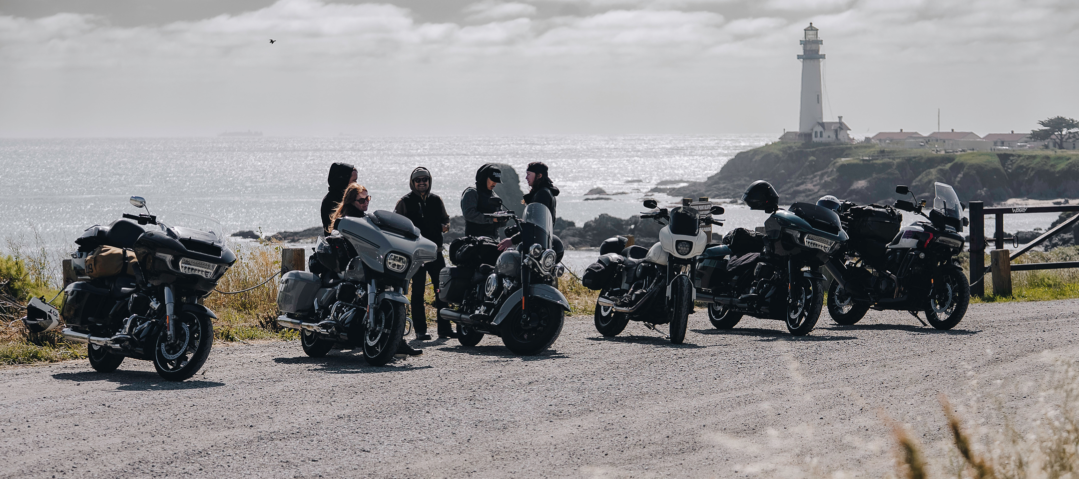 Motorcycles parked in a row at a coastal lookout with riders gathered nearby and a lighthouse overlooking the ocean