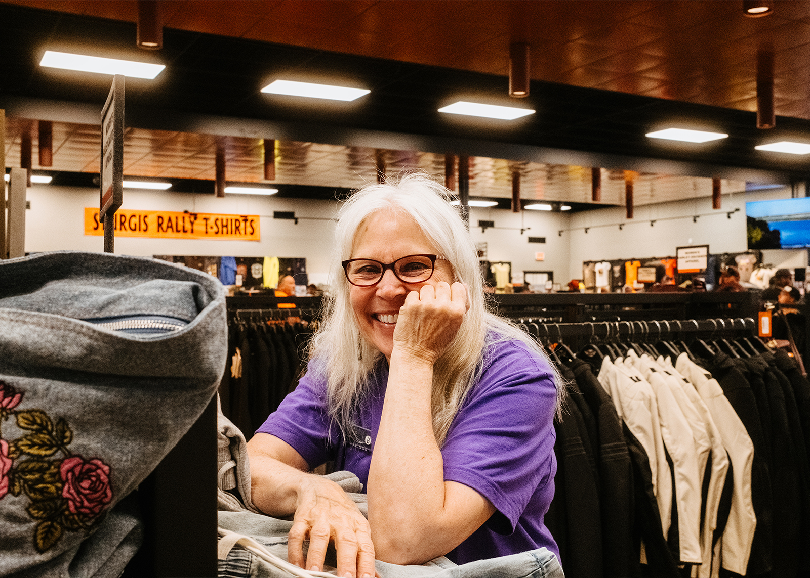 Smiling female staff member in glasses and a purple shirt inside the Sturgis Harley-Davidson motor apparel section with racks of clothing in the background.