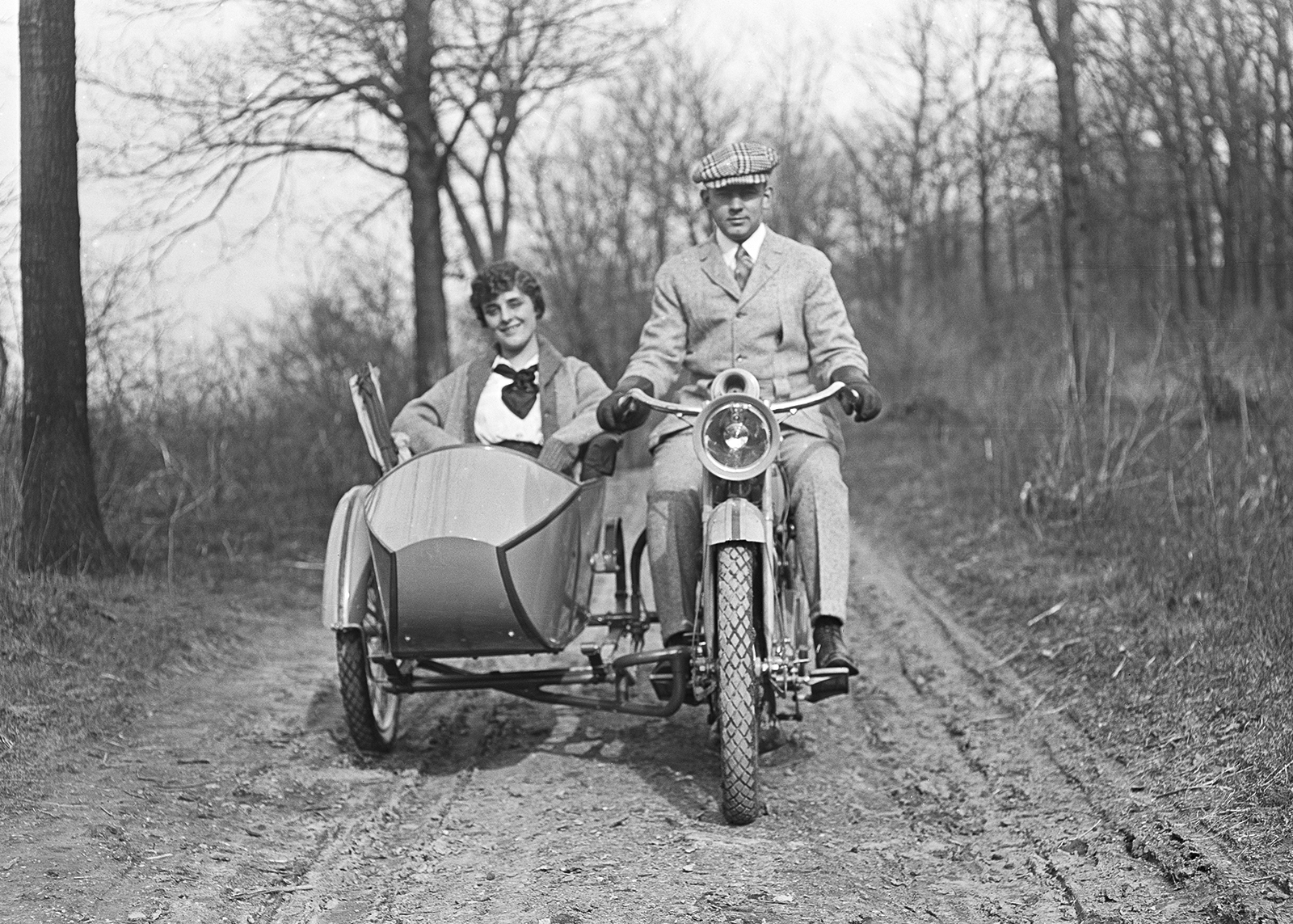 Vintage black-and-white photo of a man in suit and cap rides vintage Harley-Davidson motorcycle with smiling woman in matching sidecar.