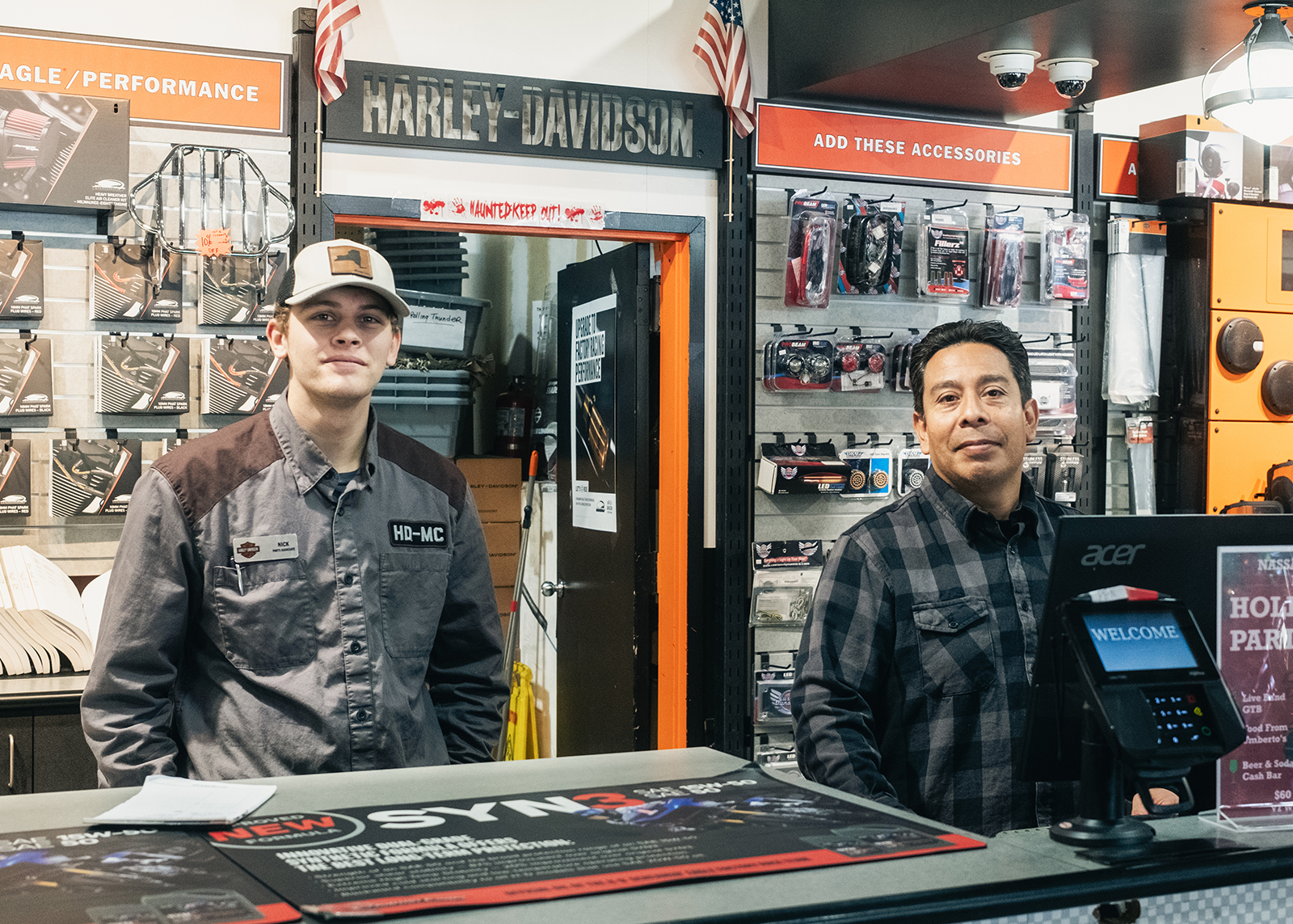 Two employees at Harley-Davidson of Nassau County stand behind a counter in front of parts and accessories.