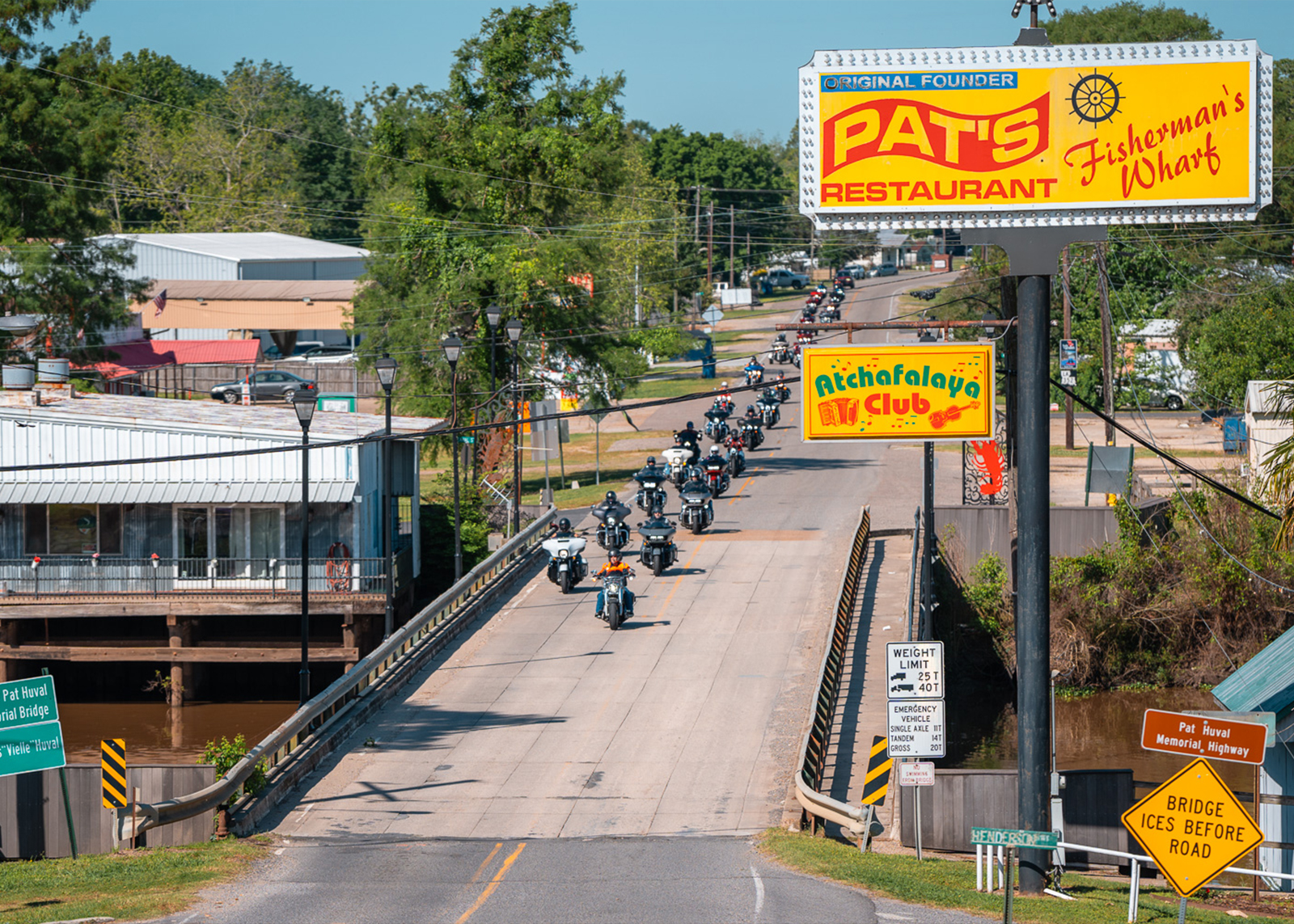 Motorcycle group riding across a bridge with bright yellow signs for Pat’s Restaurant and Atchafalaya Club in the foreground