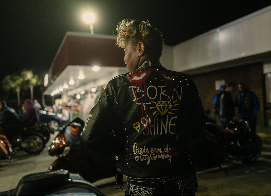 Person wearing a leather jacket with hand painted text and graphics on the back at a nighttime bike event