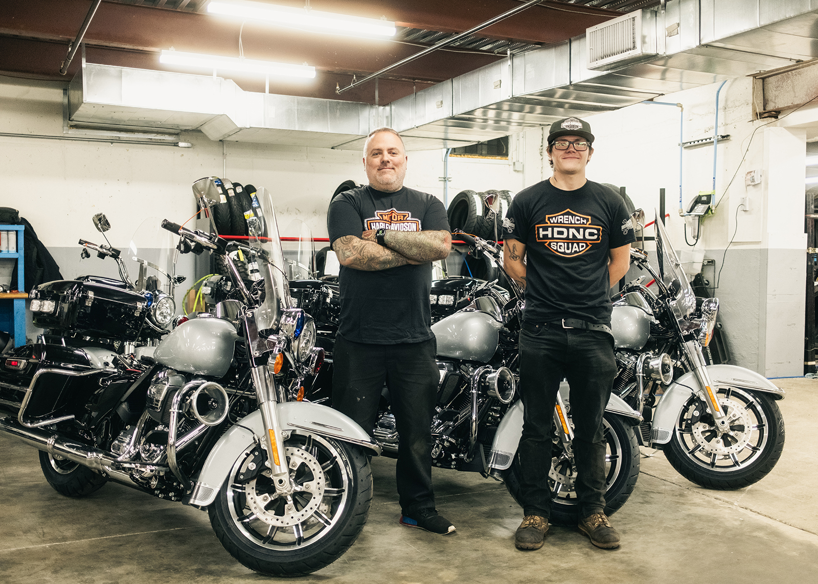 Two mechanics pose for a photo surrounded by motorcycles in the service department at Harley-Davidson of Nassau County.