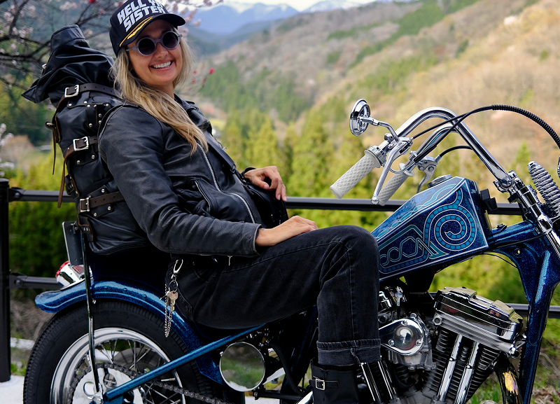 Becky Goebel seated on a custom motorcycle overlooking a mountainous landscape with greenery and a road curving in the distance