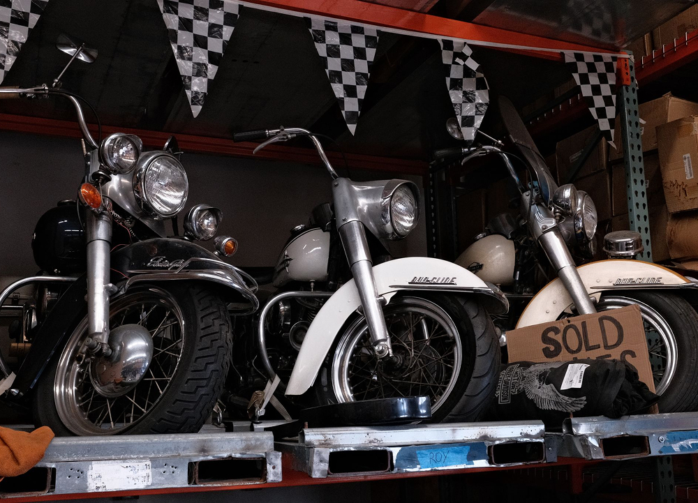 Vintage H-D motorcycles grouped on pallet with cardboard sold sign and checkered flags in workshop setting