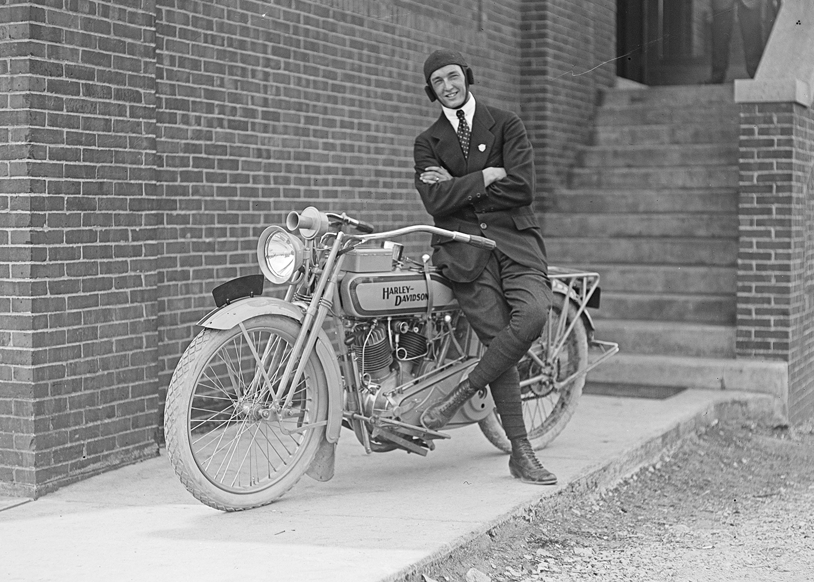 Vintage black-and-white photo of a man in suit and cap leaning on a Harley-Davidson motorcycle.