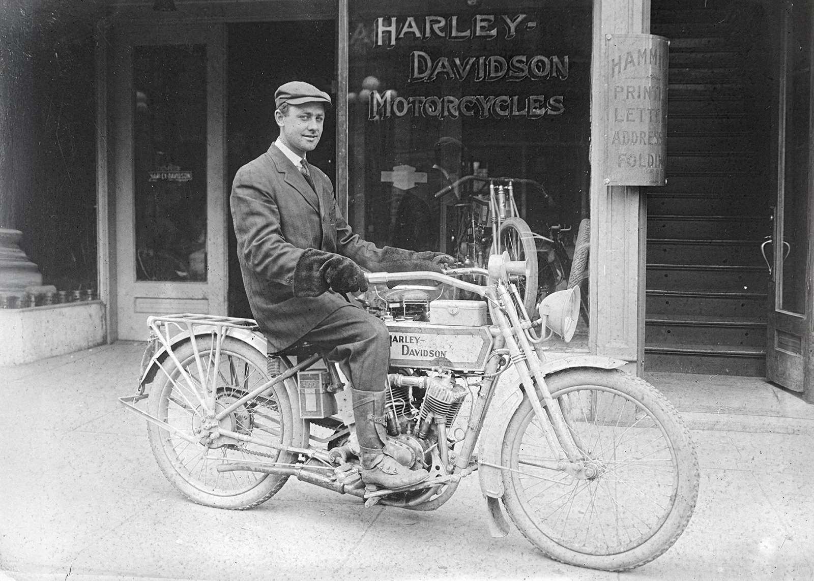 Vintage black-and-white photo of a man in suit and cap sitting on a vintage Harley-Davidson motorcycle outside a Harley-Davidson dealership.