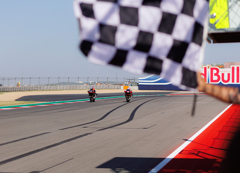 Motorcycle racers approach the finish line as a checkered flag waves overhead on the main straight