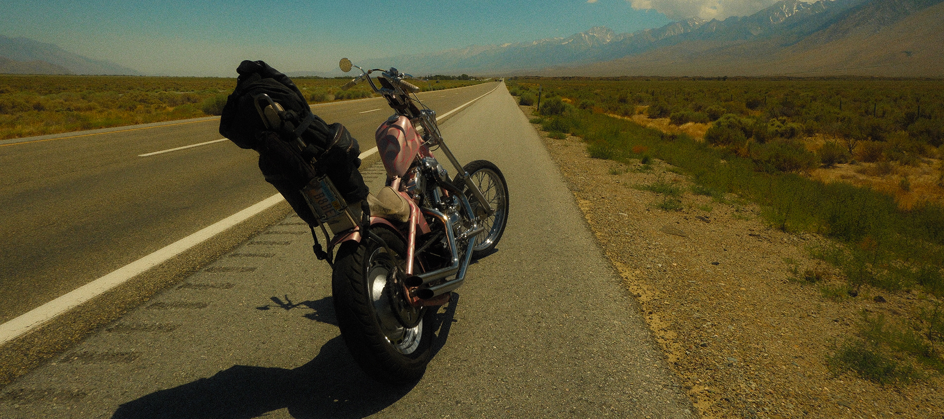 Custom chopper motorcycle loaded with travel gear parked along a desert highway with distant mountains and open landscape stretching to the horizon