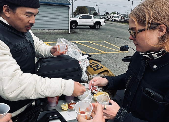 Group gathered in a parking lot sharing oysters from small cups sitting on the luggage rack of a parked motorcycle