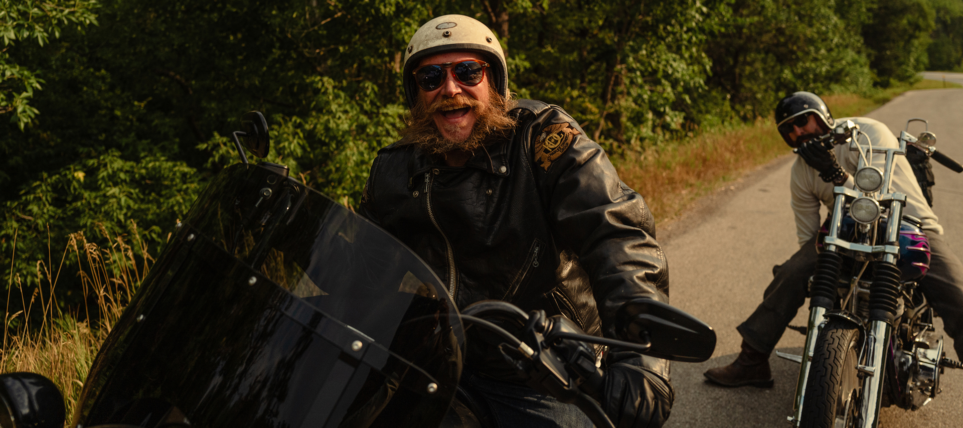 Two motorcyclists pause on a tree-lined backroad, posing with custom bikes during a scenic ride
