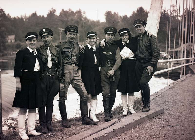 Line of riders in uniform-style outfits standing near a waterfront dock with boats behind them