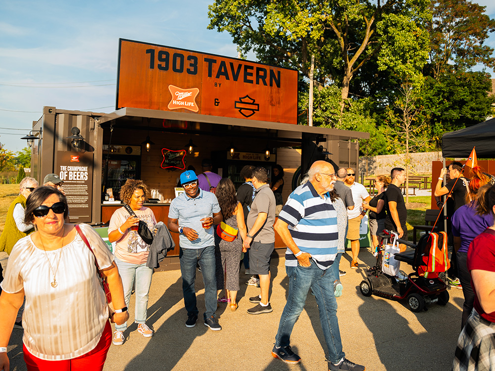 Crowd at a “1903 Tavern” stand with drinks outdoors