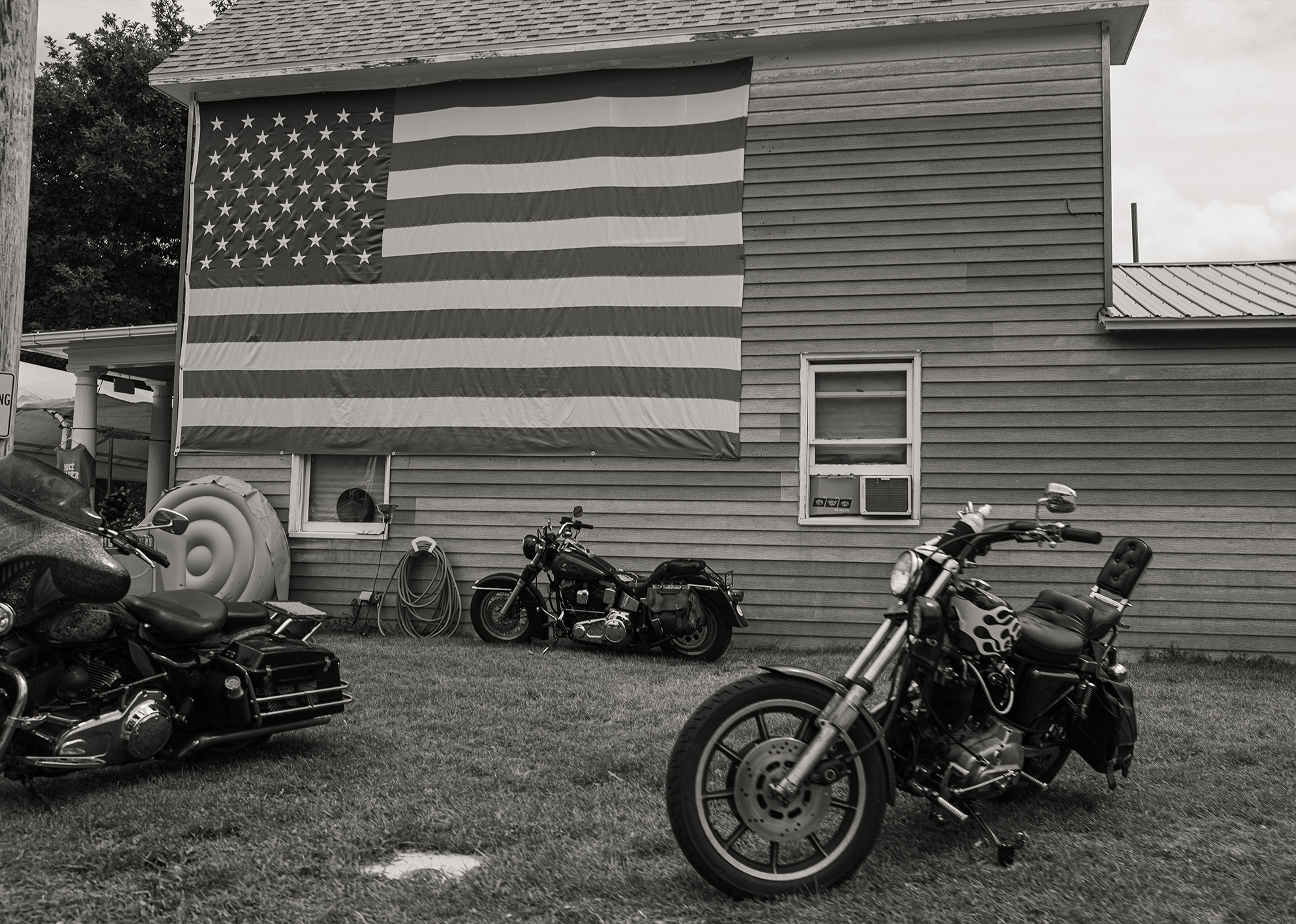 Motorcycles parked in front of a house with a large American flag hanging on the siding.