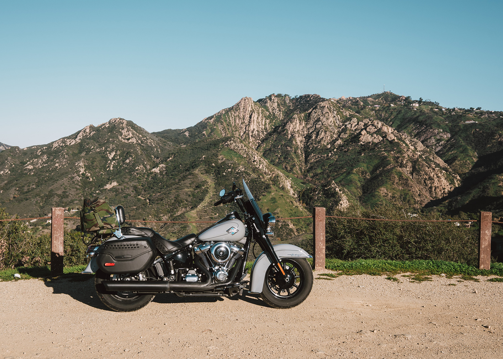 A Harley-Davison Heritage Classic motorcycle parked on a dirt roadside turnout with rugged green mountains in the background