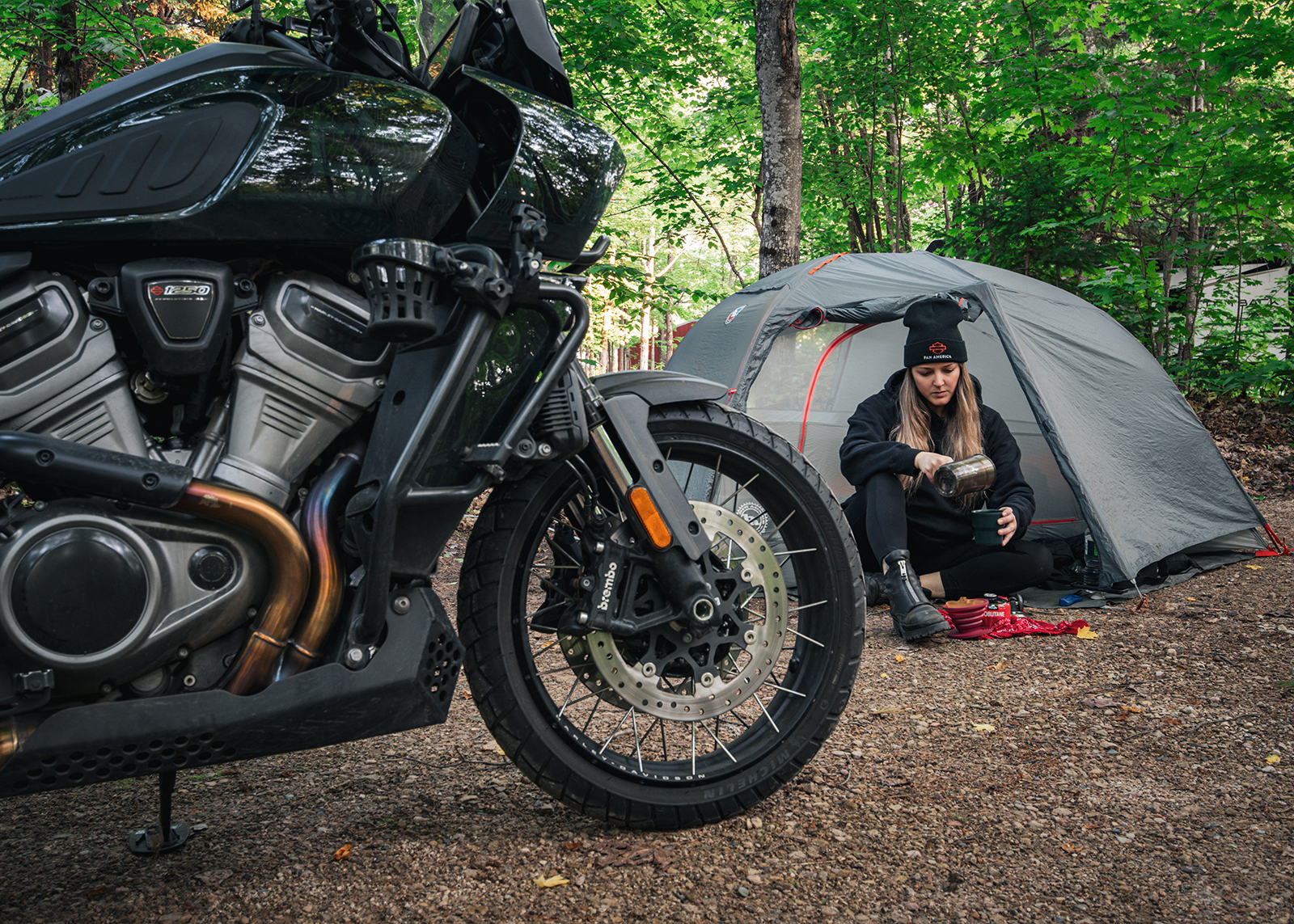 Motorcycle parked at a forest campsite as the author in a beanie pours coffee outside her tent.