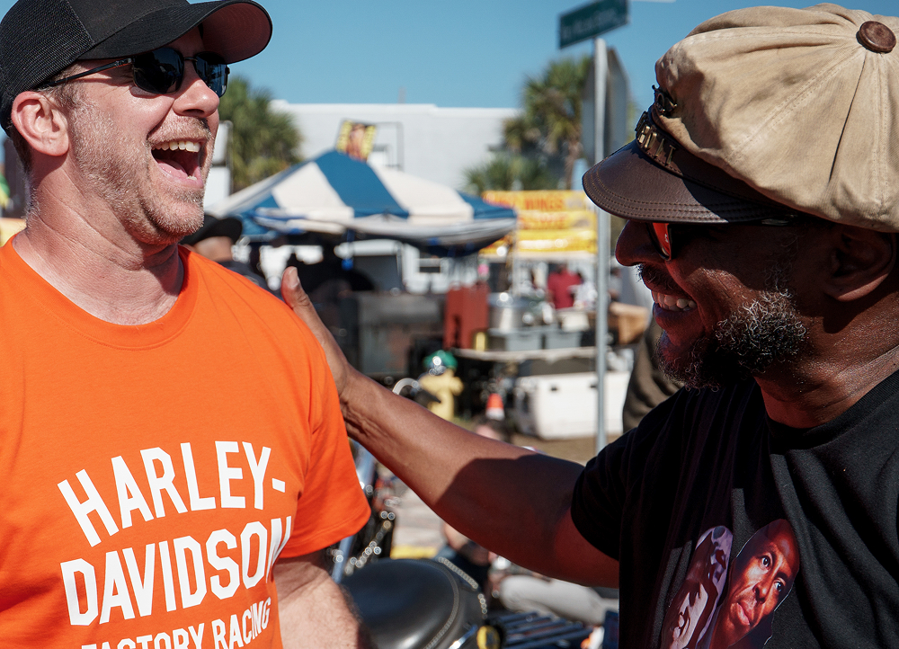 Two people laugh and talk closely at an outdoor motorcycle gathering with tents, bikes, and crowds in the background