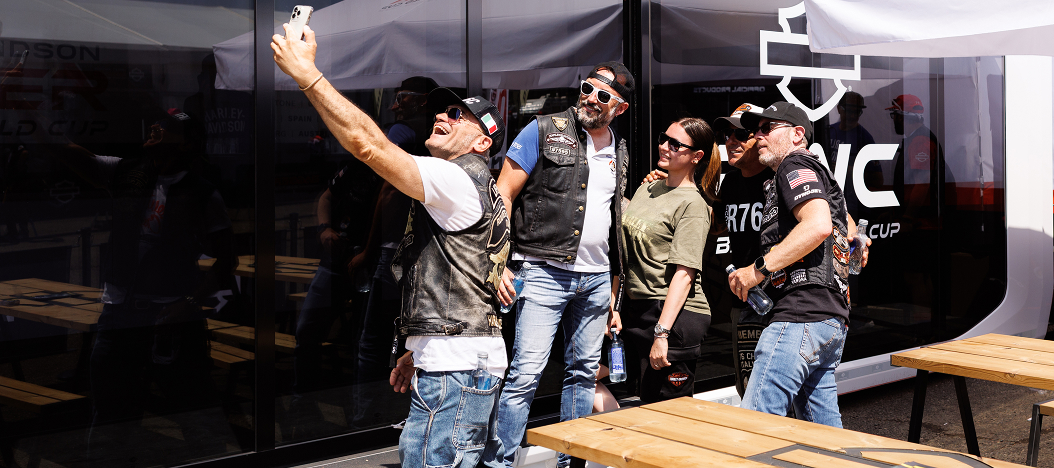 Group of Harley-Davidson enthusiasts taking a selfie outside a branded event display at a race weekend venue