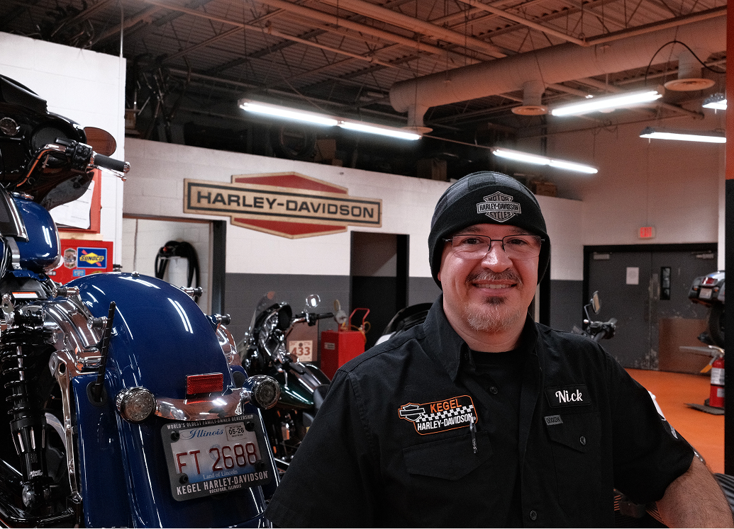 A technician stands next to a blue motorcycle on a lift in the service department at Kegel Harley-Davidson
