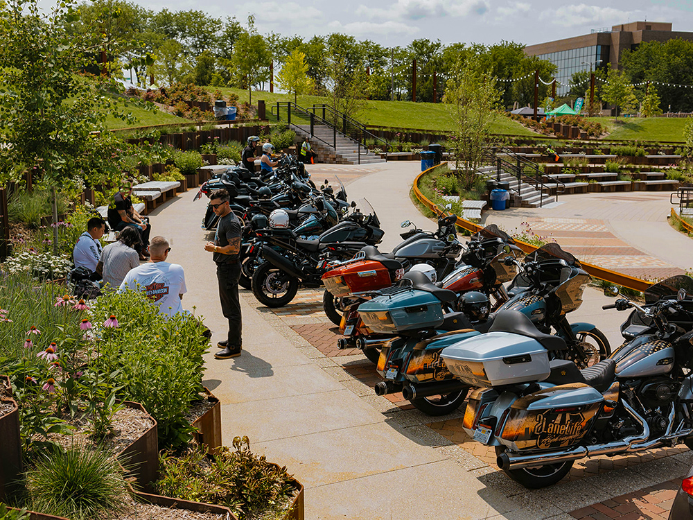 Motorcycles parked along a path with people gathered in a landscaped outdoor area