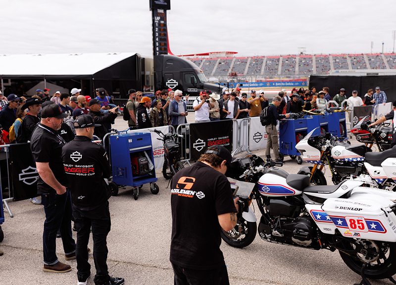 Crowded paddock scene with fans and media watching crew members line up race motorcycles
