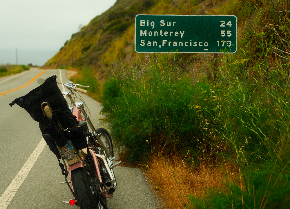 Custom chopper motorcycle parked beside a highway sign showing distances to Big Sur, Monterey, and San Francisco