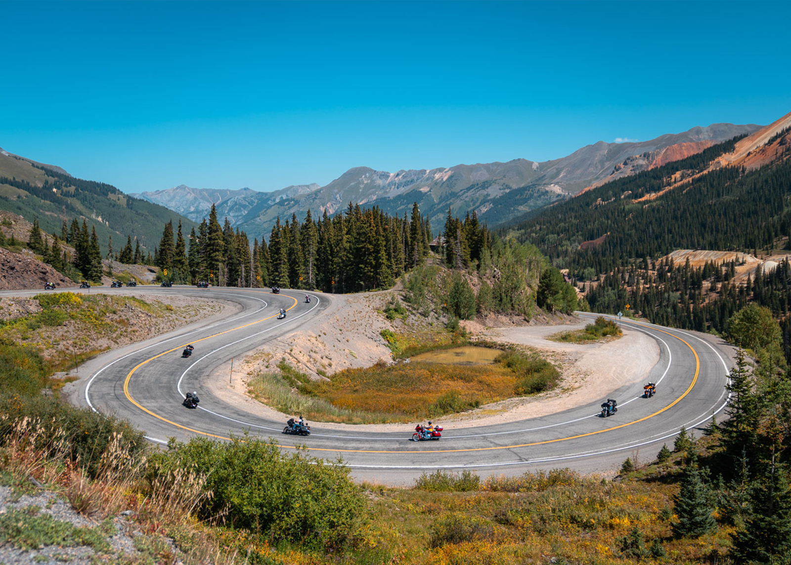 A group of motorcycles rides around a horseshoe bend curve in the mountains