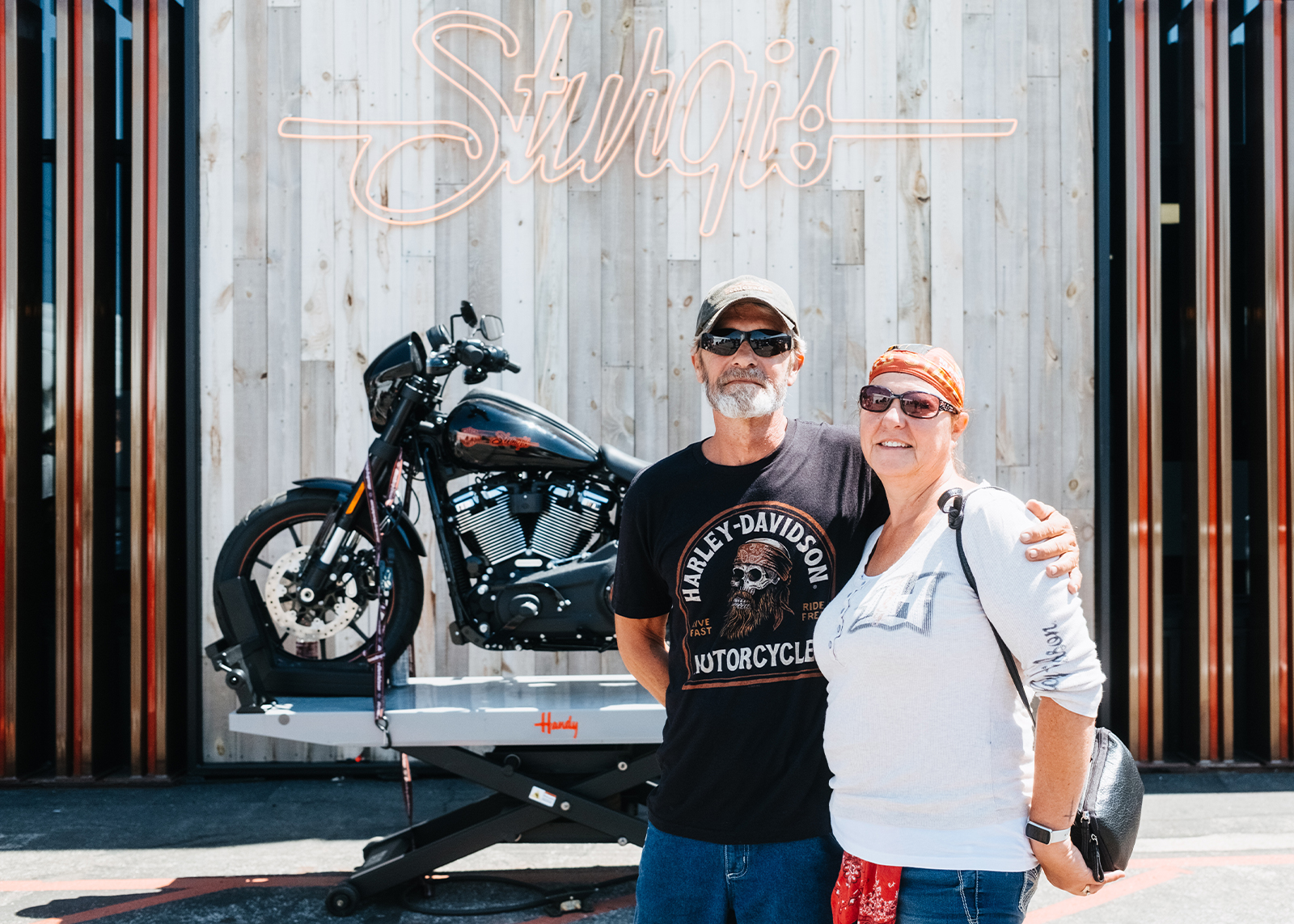 A couple in town for the Sturgis Motorcycle rally pose in front of black Harley-Davidson and neon Sturgis sign at the Sturgis Harley-Davidson dealership.