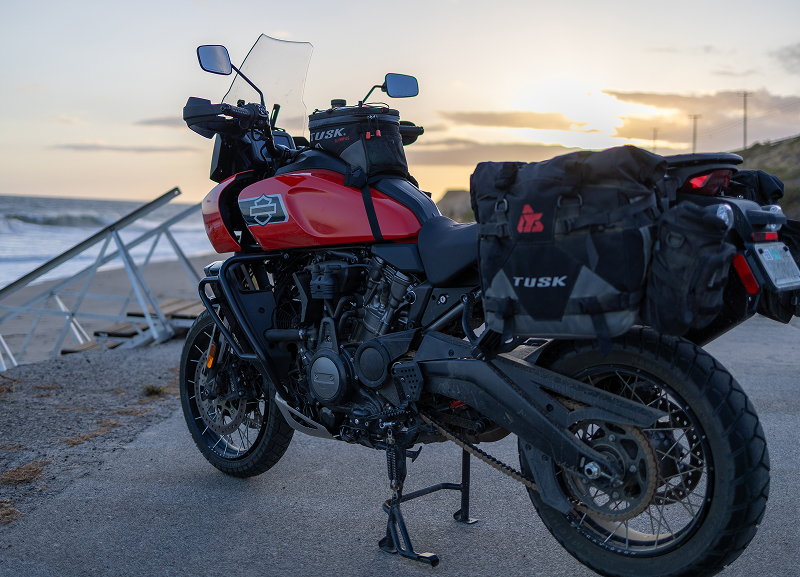 Red H-D Pan America motorcycle with luggage parked near beach access at sunset with ocean and soft sky light
