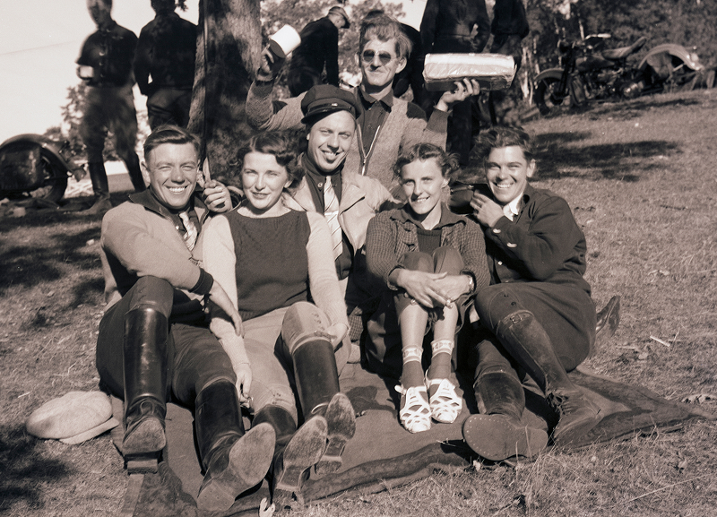 Vintage photo of group of riders sitting together on grass beside parked motorcycles, gathered casually outdoors