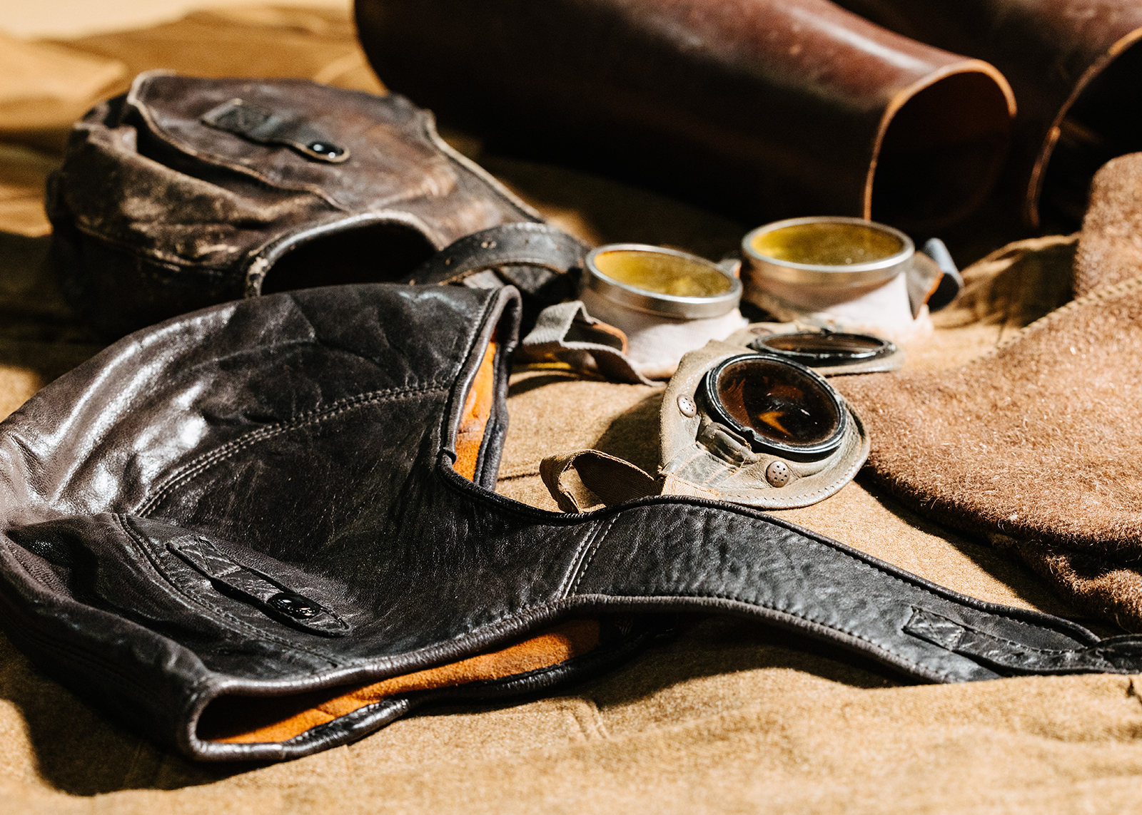 Close-up of vintage leather motorcycle helmet, gloves, and goggles with tinted lenses on brown fabric.