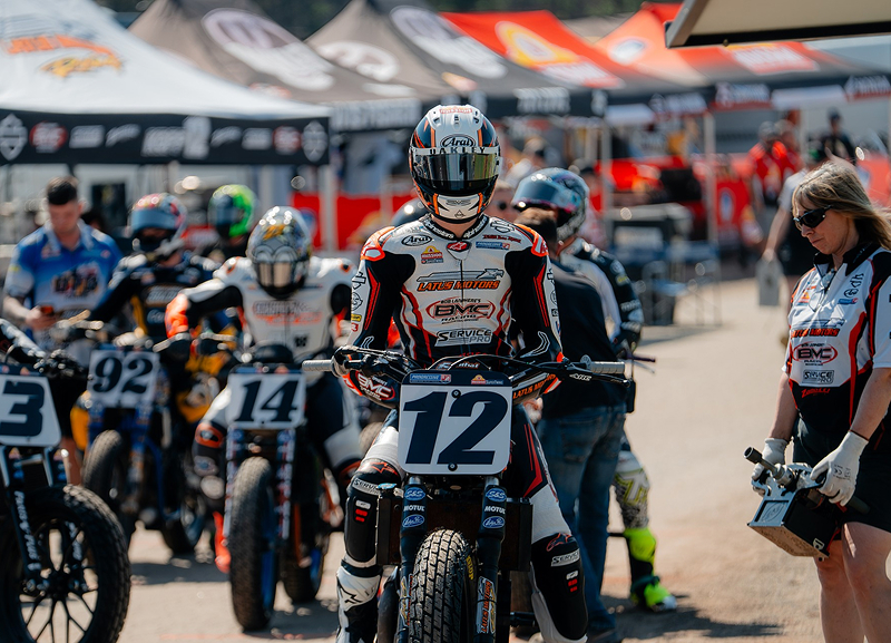 Riders roll through paddock area with number plates visible as crew members walk alongside under tents