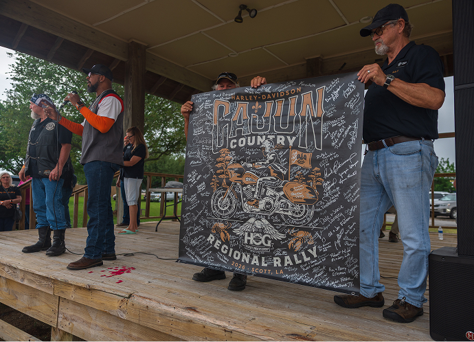 People stand on a small stage holding a large signed event banner during a Harley-Davidson regional rally