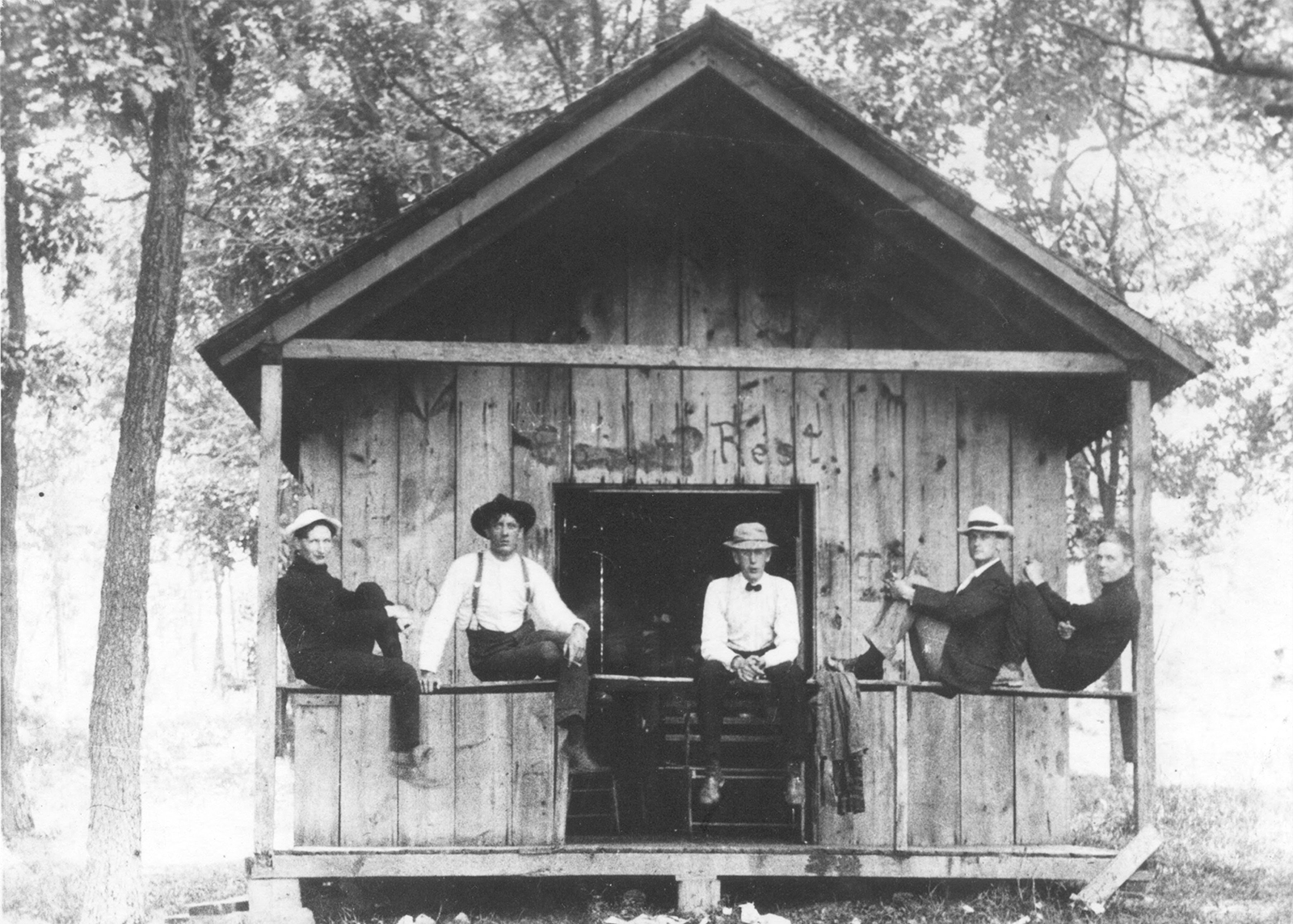 five men sit on a porch rail outside of a shed 