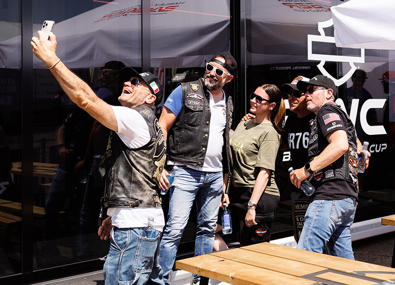 Group of Harley-Davidson enthusiasts taking a selfie outside a branded event display at a race weekend venue