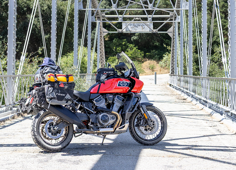 Red H-D Pan America motorcycle with full luggage parked on steel bridge with trees and shadows in bright sunlight