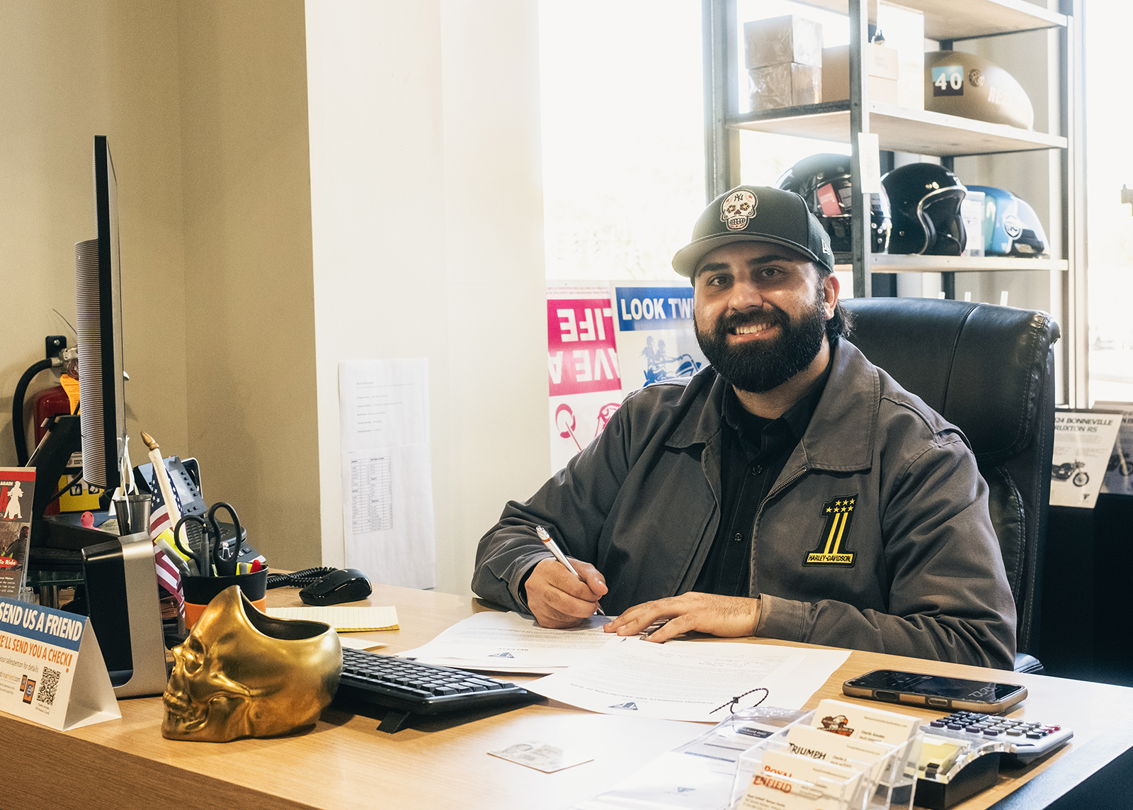 An employee at Harley-Davidson of Nassau County smiles for the camera sitting behind a desk with a pen in his hand.